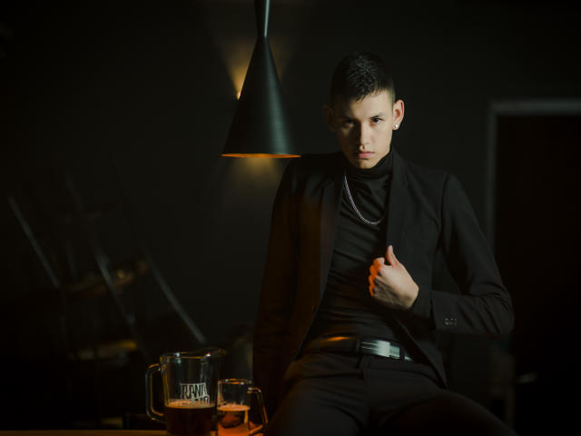 JeanKarloLeone, with short dark hair, sits under a black lamp, wearing a black turtleneck and suit, right hand on chest, looking serious, with a glass of dark beer.