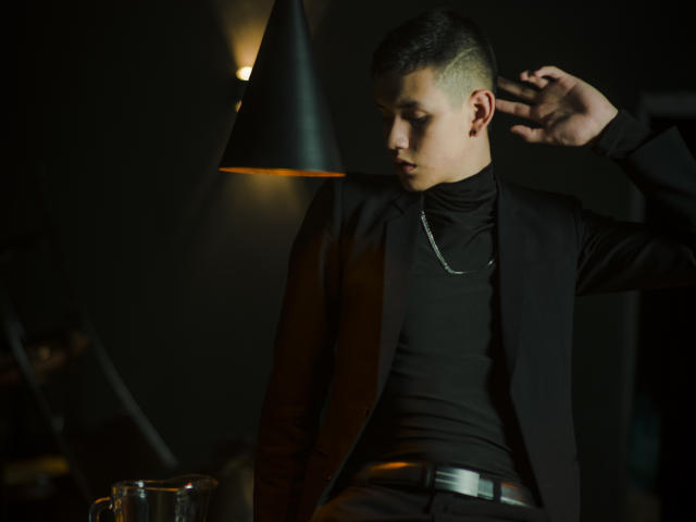 JeanKarloLeone with short, buzzed hair, wearing a black turtleneck, suit, and chain, under a black lamp, touching his ear, with a glass on a table.