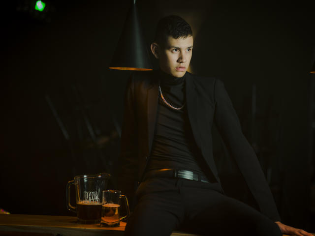 JeanKarloLeone, short black hair, black suit, black turtleneck, black pants, sitting on wooden bar, black lamp overhead, beer mug with "craft beer" on left, dark background, serious expression.