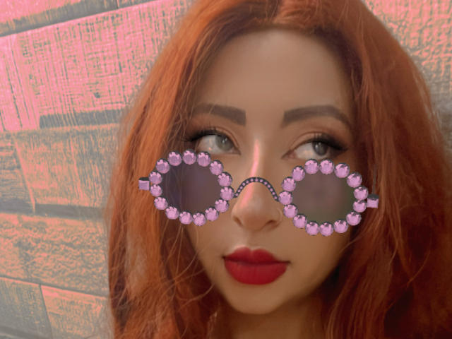 RavenAndEllie with red hair, wearing pink gemstone sunglasses, and red lipstick, looking up with green eyes. Her ass is not visible. The focus is on her face.