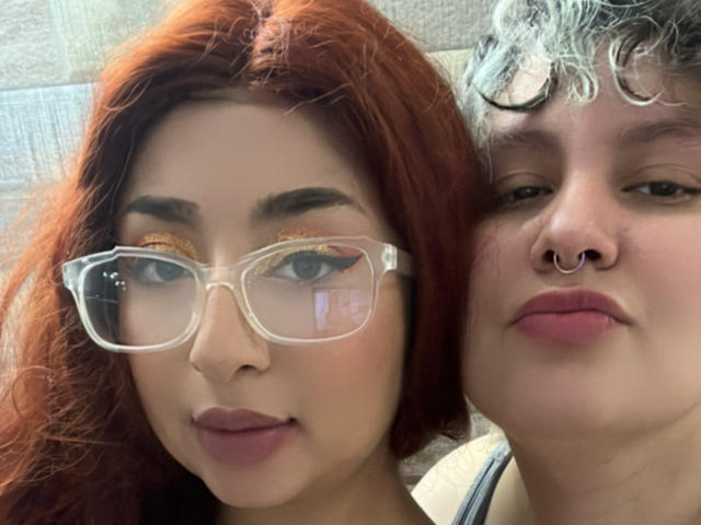 RavenAndEllie with red hair, glasses, and pink lipstick, next to Ellie with curly gray hair, nose ring, and pink lipstick. Both have soft smiles and relaxed expressions.