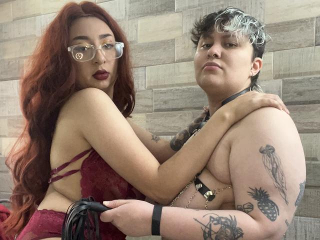 RavenAndEllie with red hair, glasses, and dark lipstick, holding a black leash, arm around Ellie with short, gray-tipped hair, tattoos, and a black harness.