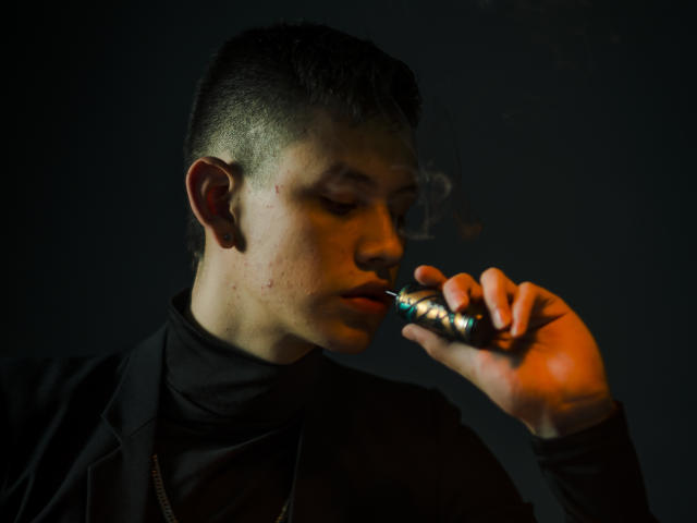 JeanKarloLeone with a cigarette, holding it near his mouth, wearing a black turtleneck and jacket, short dark hair, smoky breath, dark background, focused expression, slightly dirty skin.