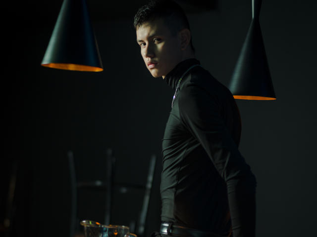 JeanKarloLeone with short dark hair, wearing a black turtleneck, looking over shoulder, under dark green cone-shaped lights, serious expression, dark background, hands by side.