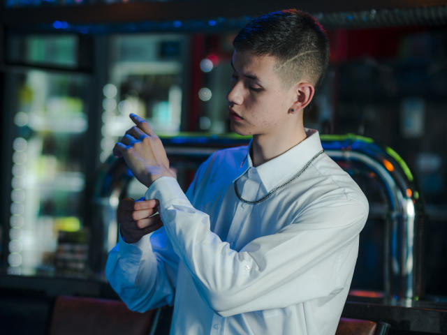 JeanKarloLeone, wearing a white shirt and silver chain, adjusts his wrist in a bar with blue and red lighting, looking focused with short, styled brown hair.