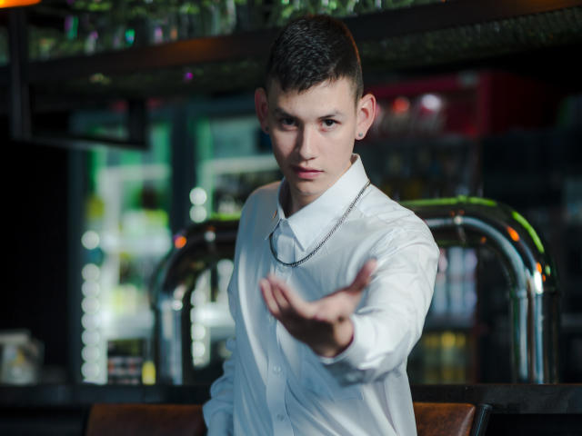 JeanKarloLeone, with short brown hair and white shirt, extends his right hand forward, looking serious. He's wearing a silver chain, standing in a dimly lit bar.