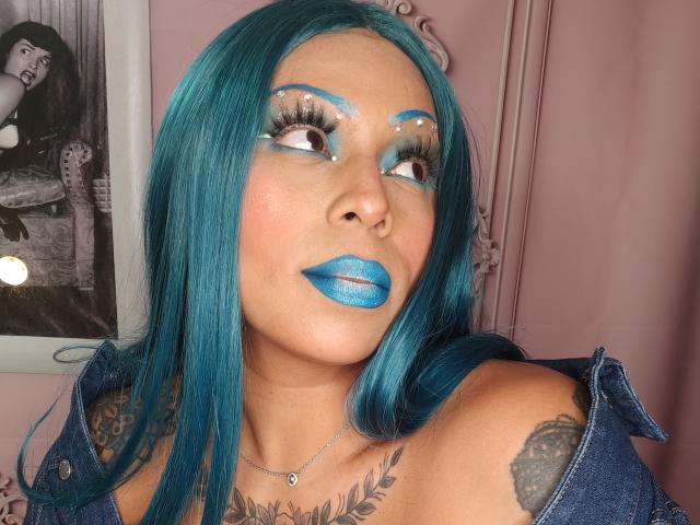 Chili with teal hair and blue lipstick, looking upward, has tattoos on her shoulders, wearing denim top, glowing makeup.