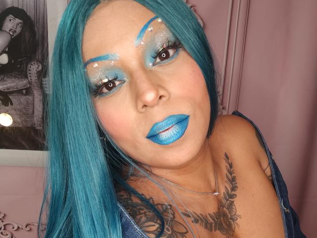 Chili, with bright blue lipstick and matching eyeshadow, faces the camera, her long blue hair covering her shoulders, looking sexy.