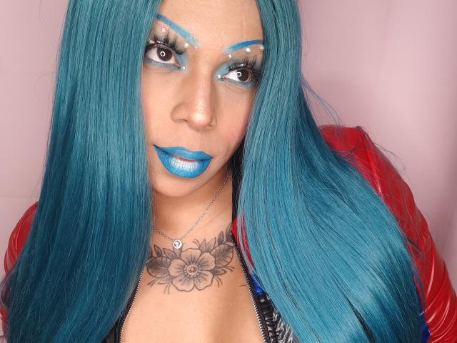 Chili with bright blue lipstick and matching hair, wearing a red jacket, looking up seductively, floral tattoo around neck.