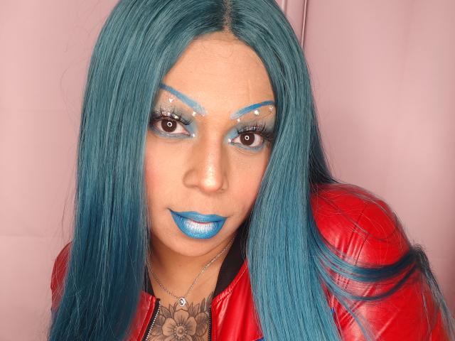 Chili with long blue hair and blue lipstick, wearing red leather jacket, has blue eyeshadow and makeup, looking at camera.
