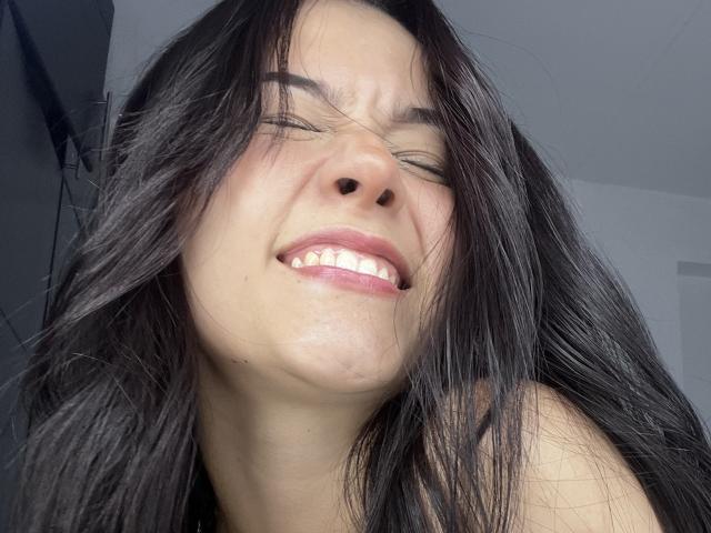 DeisyWest with black hair, eyes closed, smiling with teeth showing, looking slightly annoyed, face squinting, light skin, no clothes visible, background blurred, photo.