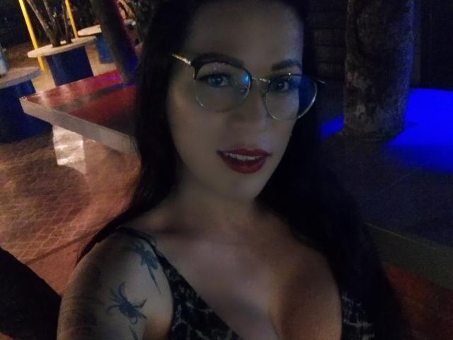 SexyCaandydoll with glasses, red lipstick, black hair, tattoos on her right shoulder, and a low-cut black dress, showing her tits in a dimly lit room.