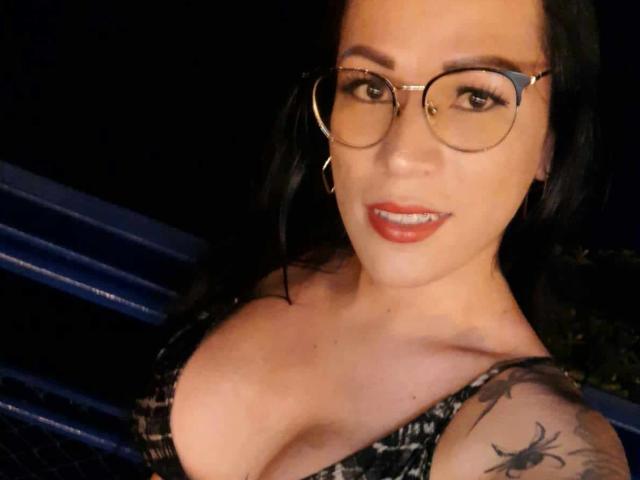 SexyCaandydoll with glasses, red lipstick, black hair, and tattoos, showing her boobs in a low-cut top, smiling at the camera.