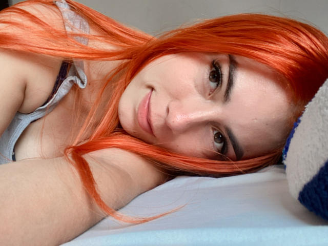 JuliaGarciax with orange hair, lying down, head on pillow, smiling, brown eyes, wearing a grey top, visible cleavage, soft light on her face, relaxed, cute.
