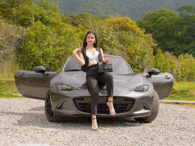 RacheFoxx sits on a gray sports car with doors open, wearing a white crop top, black pants, and heels, holding a black handbag, showing her tits and ass.