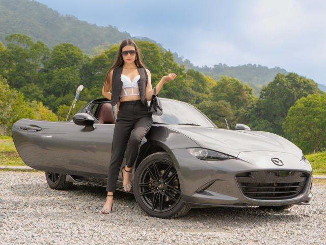 RacheFoxx with long brown hair, sunglasses, white bra, black pants, and heels, standing next to a gray Lotus Elise with black wheels and gravel driveway.