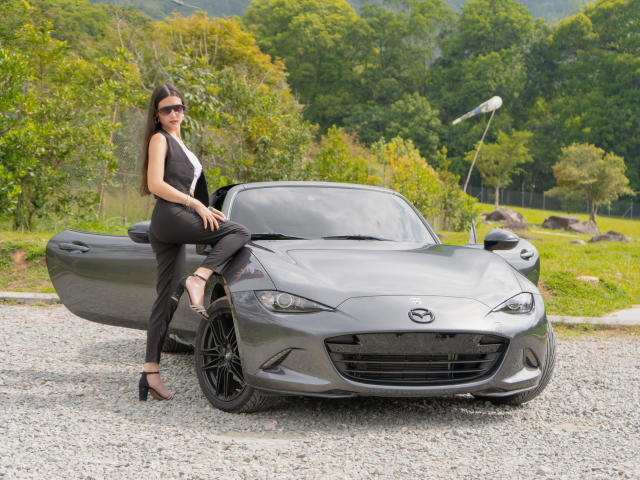 RacheFoxx, with her ass in black pants and high heels, sits on a gray Nissan sports car, showing her tits in a tight black top.