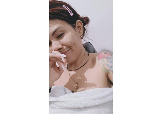 NathashaRomexo a la peau brune, les cheveux roux et une pince à cheveux rose. Elle sourit, avec de longs ongles, un collier et un tatouage coloré sur son épaule, enveloppée dans une serviette blanche..