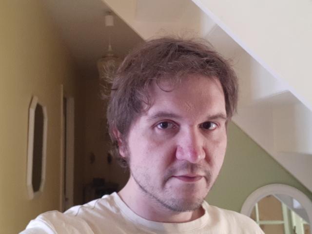 NectarineFad has a chubby face with curly brown hair, a light beard, and is wearing a white shirt. His nose is slightly red. He's looking directly at the camera.