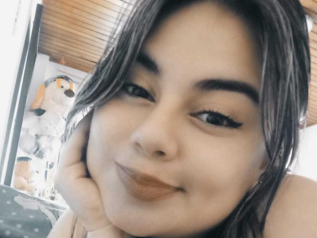 ZafiroLeroy has medium brown skin, black eyebrows, long eyelashes, and brown lipstick. She's smiling with her hand on her cheek, brown hair, and a stuffed dog in the background.