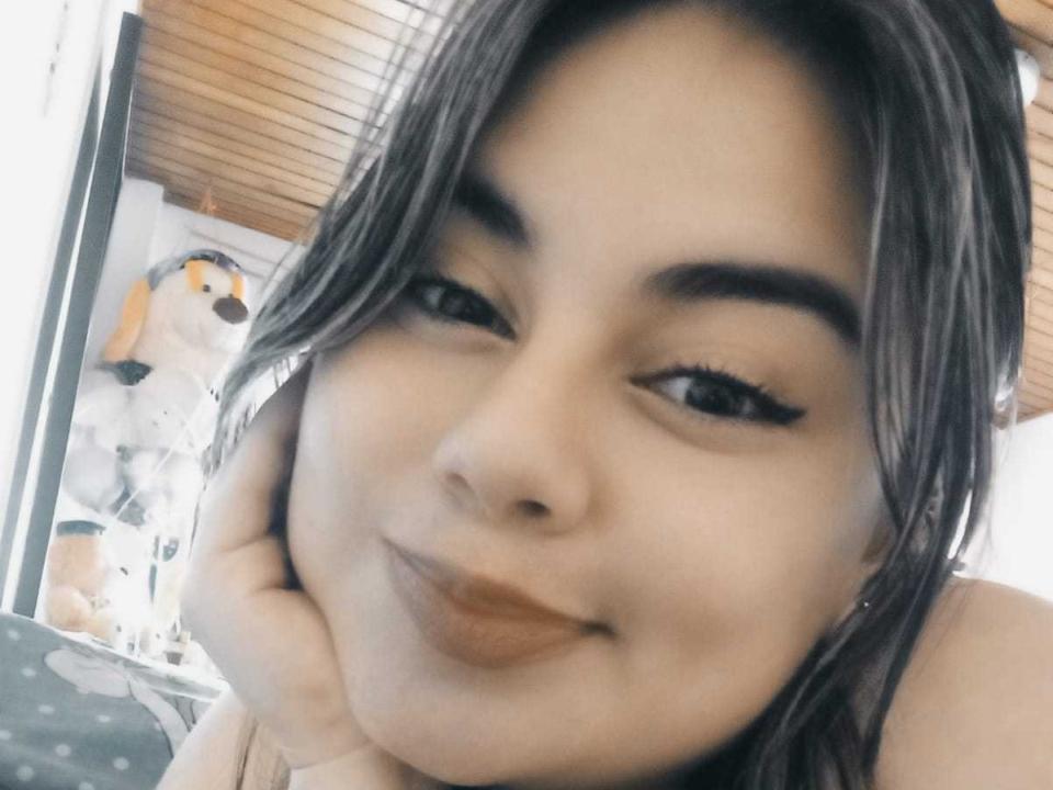 ZafiroLeroy has medium brown skin, black eyebrows, long eyelashes, and brown lipstick. She's smiling with her hand on her cheek, brown hair, and a stuffed dog in the background.