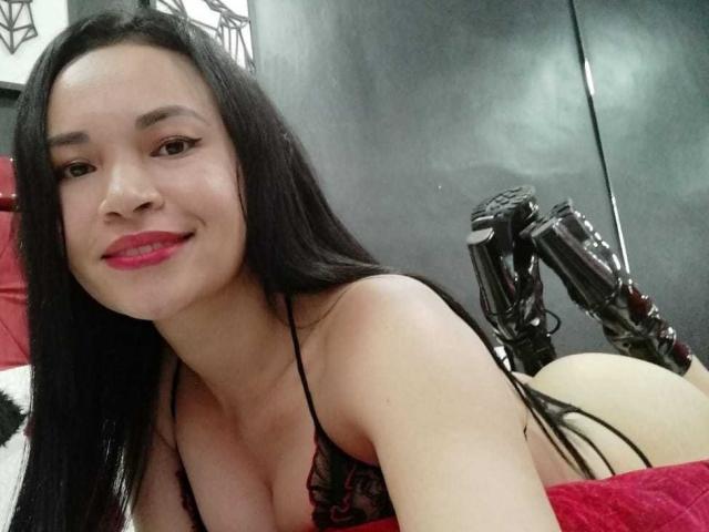 AiconWarrior with long black hair, red lipstick, and black lace bra, lying on bed, smiling, showing her tits and ass, with black boots in the background.