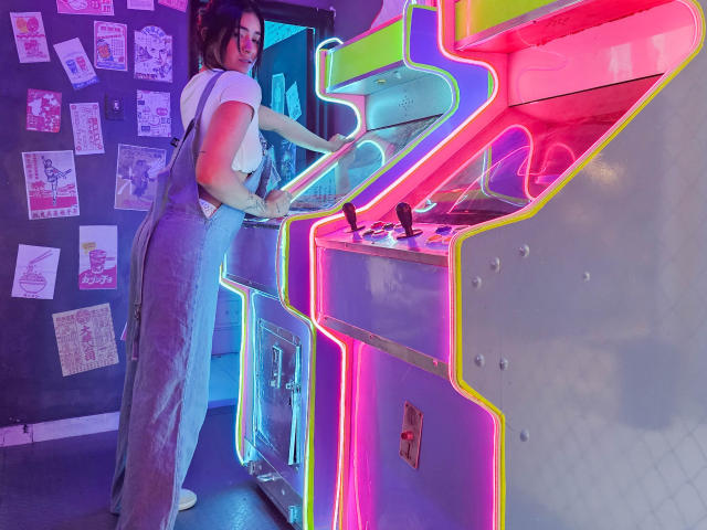 NicoColeman with tits in a white shirt, denim overalls, and glasses, playing a neon-lit arcade game, standing with ass slightly bent, hands on the machine.