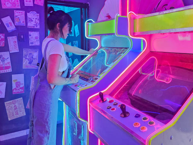 NicoColeman with tattoos, in a white tee and blue apron, playing a neon-lit arcade game, her hand on the buttons, standing next to a colorful machine.
