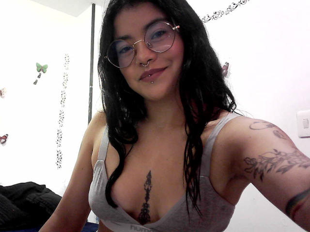 EmmaRuiz with black hair, glasses, nose ring, and lip piercing, wearing a gray bra, showing tattoos on her chest and arm, and a visible nipple piercing.