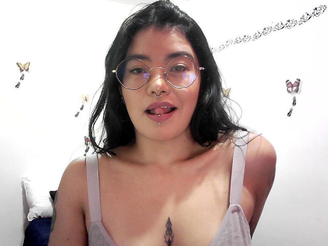 EmmaRuiz with black hair, glasses, nose ring, tongue piercing, lip piercing, grey tank top, showing small tits, butterfly wall decor, and a tattoo under her tits.