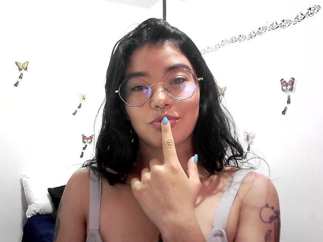 EmmaRuiz with black hair, glasses, nose ring, and blue nails, pointing at her lips, wearing a grey top, showing a tattoo on her right arm, in a selfie.