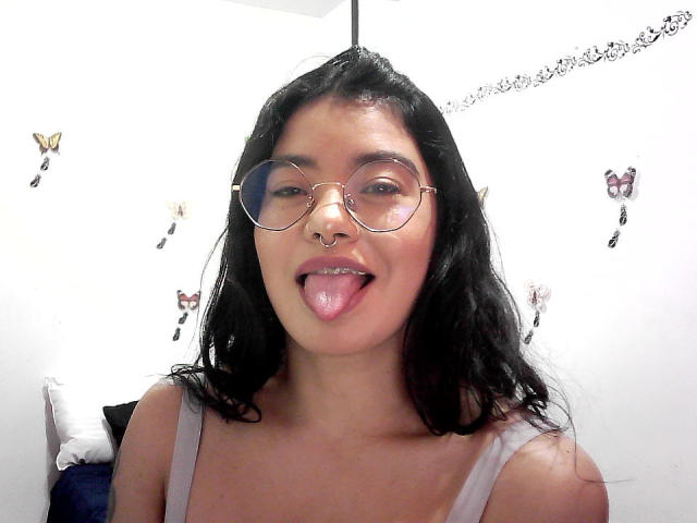 EmmaRuiz with black wavy hair, round glasses, nose ring, and tongue out, wearing a gray tank top, showing her tits. White wall with butterfly stickers in background.