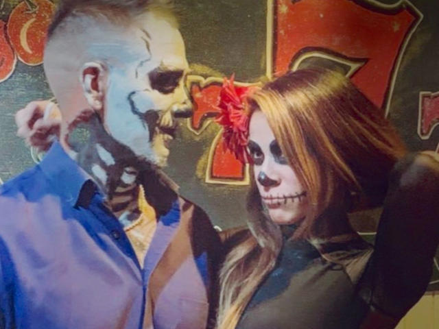 BigJLila with skeletal face paint and blue shirt, holding BigJLila's blonde-haired, red flower-crowned, black-dressed skull-faced partner, both showing grinning skull faces.