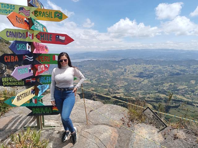 BellaSmithX69 with tits in a striped shirt, blue jeans, and sunglasses, standing on rocky mountain, holding a cane, surrounded by colorful signposts, under a blue sky.