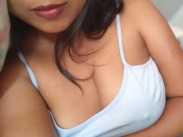 AlexiaCool, met donkerbruin haar, draagt een lichtblauw tanktopje, waardoor haar decolleté en kleine borsten zichtbaar zijn. Haar huid is bruin en haar lippen zijn glanzend roze..