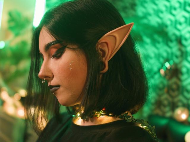 DixiPixi, com orelhas pontudas de elfo, batom escuro e cabelo preto curto, usando um colar de corrente dourada, olhando para baixo, em um quarto iluminado por luz verde, com maquiagem discreta e sardas..