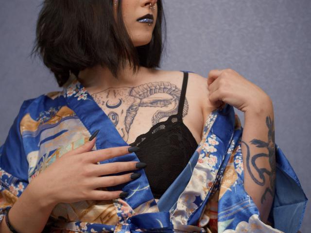 DixiPixi with black lipstick, blue nails, and a blue floral kimono, holding open to reveal black lace bra, showcasing tattoos on chest and arm. Boob tattoos visible.