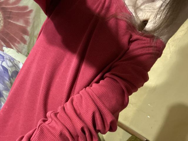 Kabblasha porte une chemise rouge à manches longues avec des côtes, ses seins étant légèrement visibles à travers le tissu, ses bras levés, et ses cheveux bruns partiellement cadrés..