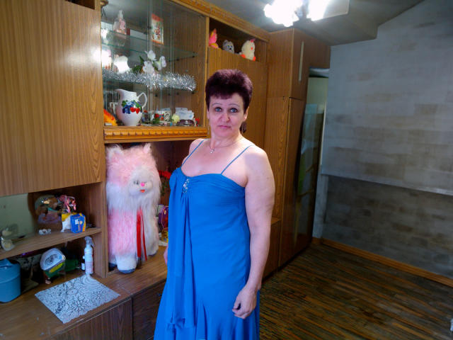 PaulaGoldens stands in a room wearing a blue dress, showing her tits, with short dark hair, a neutral expression, and a fluffy pink cat statue to her left.