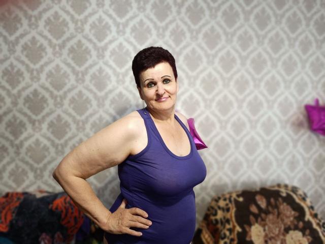 PaulaGoldens with short dark hair, smiling, wearing a purple tank top that shows her big boobs, standing with one hand on her hip, in a cozy, patterned room.