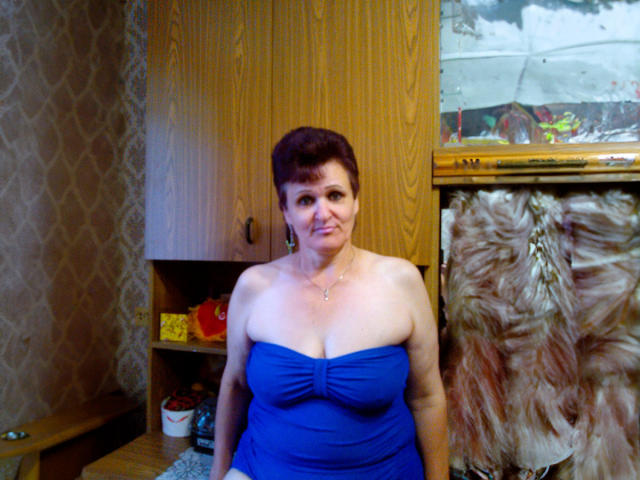 PaulaGoldens with short brown hair, wearing a blue strapless dress, showing her big boobs and cleavage, standing in a wooden room, looking at the camera.