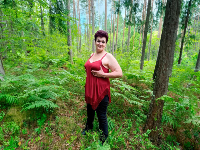 PaulaGoldens, chubby with short red hair, wearing a red, sleeveless, frilled top and black pants, stands in a green forest, smiling, holding her belly.