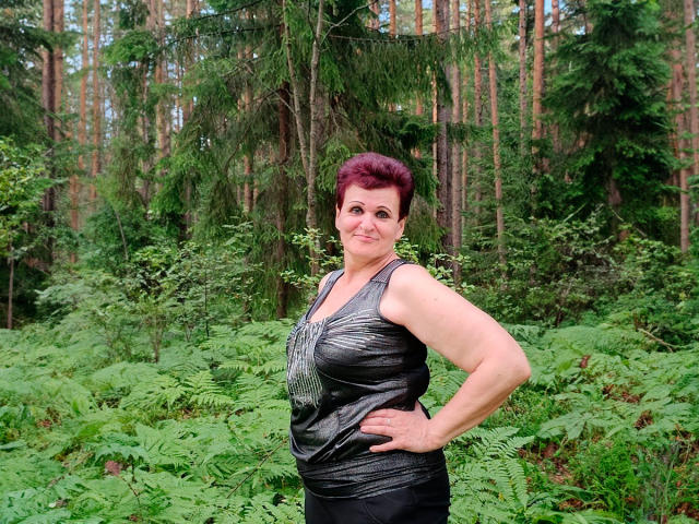 Paula, une femme aux cheveux roux, souriante, les mains sur les hanches, portant un haut noir brillant sans manches, montrant ses fesses et ses seins, se tient dans une forêt avec des fougères vertes..