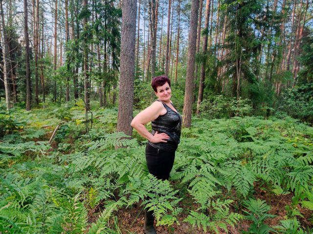 PaulaGoldens stands in a forest, wearing a black leather sleeveless top, hands on hips, smiling, with short red hair, curvy with noticeable ass, surrounded by green ferns.