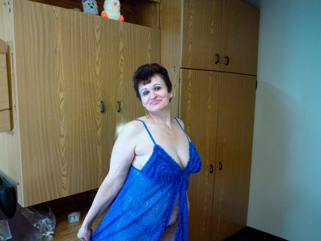 PaulaGoldens has big tits in a blue lace babydoll, smiling, with short brown hair, standing in a room with wooden cabinets, and a stuffed toy on top.