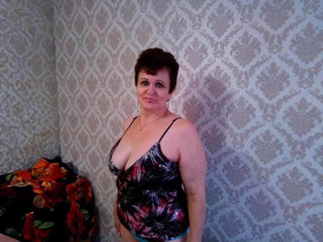 PaulaGoldens with short brown hair, wearing a black and pink floral tank top, showing her big tits and slightly smiling, standing in front of a floral-patterned wall.