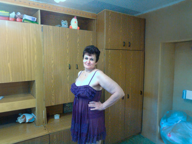 PaulaGoldens, standing in a room, wearing a sheer purple dress, showing her tits, short dark hair, smiling, hands on hips, wooden cabinets in the background.
