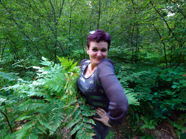 PaulaGoldens with short purple hair, wearing a dark long-sleeve top, standing in a green forest, hands on hips, smiling, holding fern leaves, showing her boobs slightly.