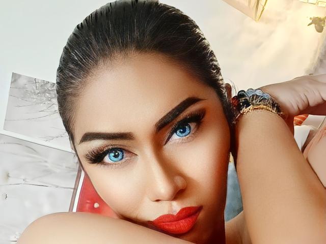 Das Gesicht von MaddisonAderson mit leuchtend blauen Augen, dicken Augenbrauen, rotem Lippenstift und zurückgekämmtem Haar, sie trägt ein Armband und blickt verführerisch in die Kamera. Ihre Brüste sind leicht sichtbar..