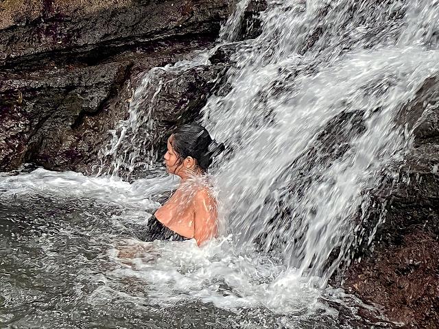 MaddisonAderson, con i capelli neri bagnati, le spalle scoperte e piccoli seni, in piedi sotto una cascata, con l'acqua che le scorre sulla schiena, una scogliera rocciosa alla destra e schizzi d'acqua..